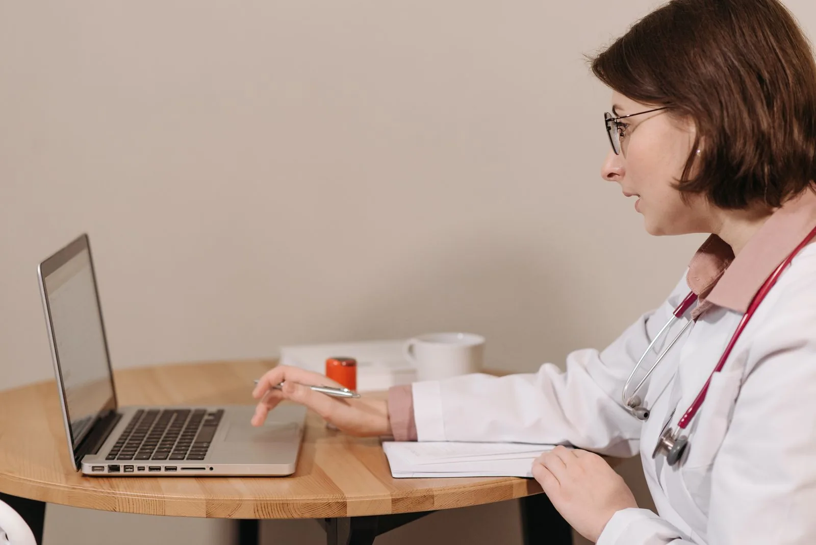 Una dottoressa in camice bianco che usa un laptop mentre scrive appunti su un tavolo di legno.