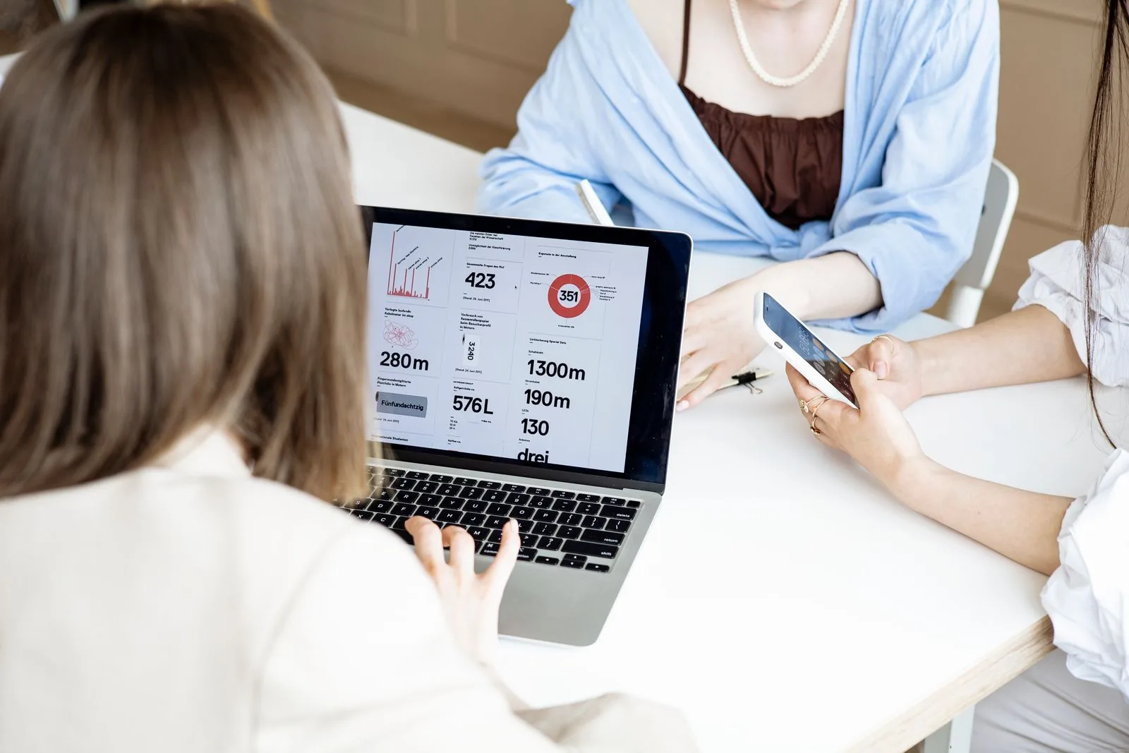 Un gruppo di donne che collaborano a una scrivania utilizzando un laptop e uno smartphone per attività lavorative al chiuso.